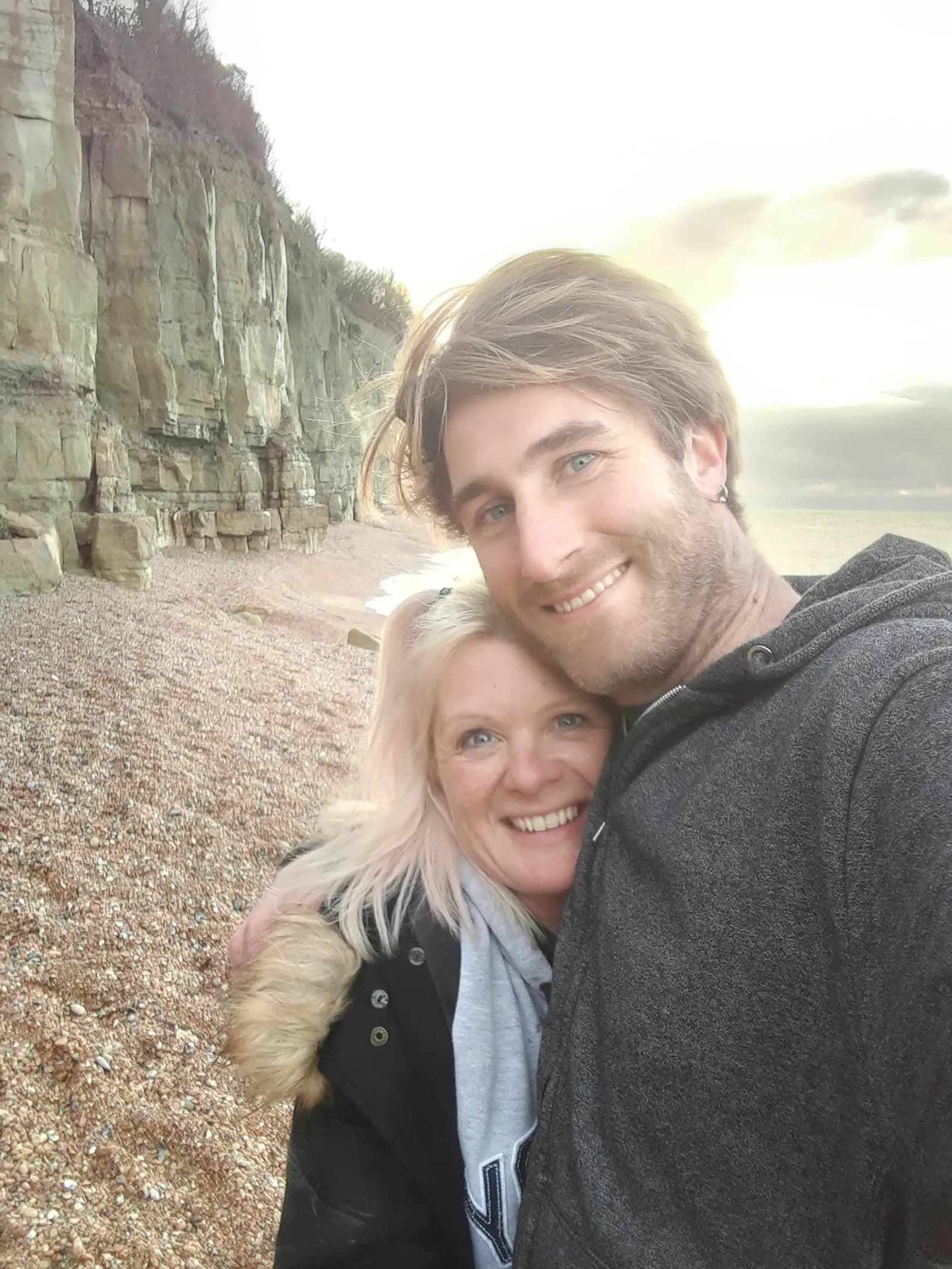 A man and a woman smile at the camera on a pebbly beach with cliffs and the ocean in the background.